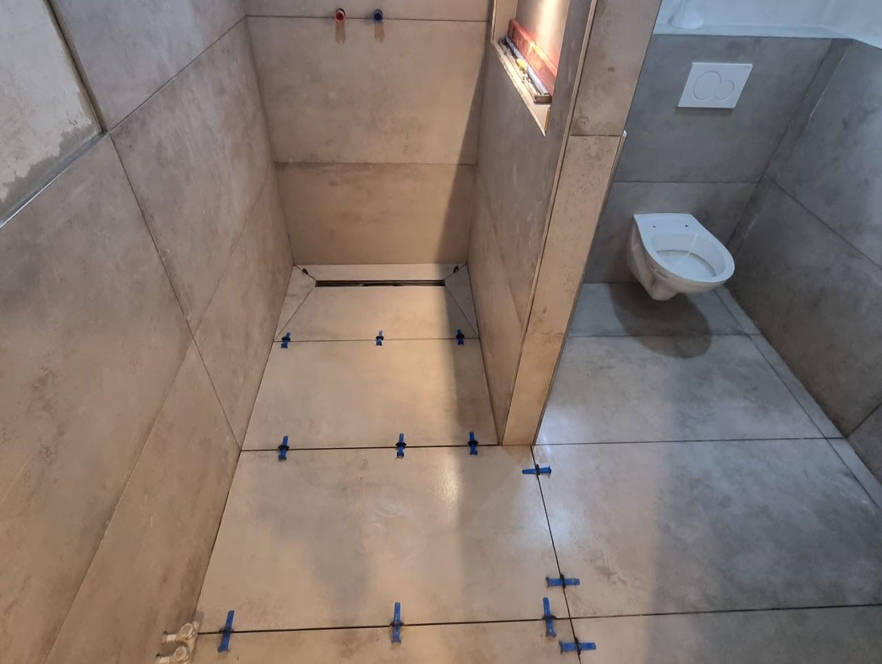 Das Badezimmer wird renoviert und hat graue Fliesen an Wänden und Boden. Toilette und Abfluss sind sichtbar.