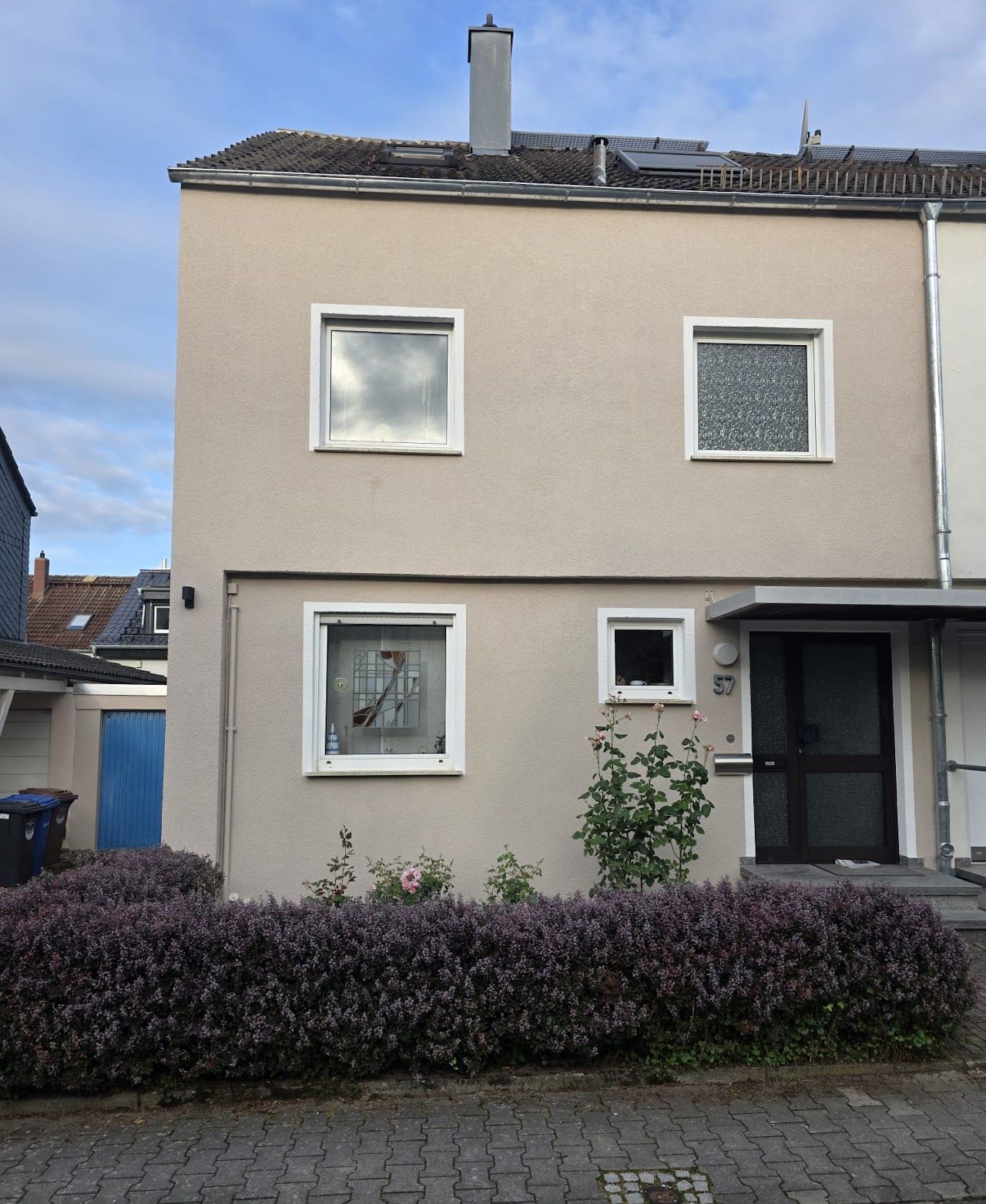 Zweistöckiges beigefarbenes Gebäude mit Fenstern und einer dunklen Tür, davor kleine Pflanzen und eine violette Hecke.