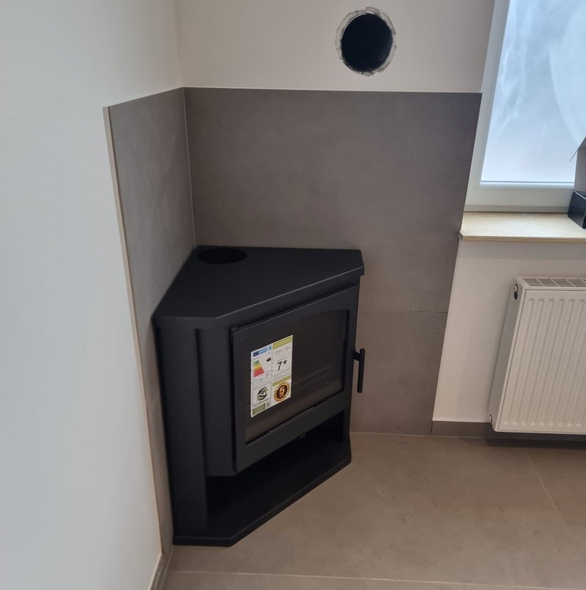 Schwarzer Eckofen in einem Zimmer, unter einem Lüftungsloch, neben einem Fenster und einem Heizkörper.