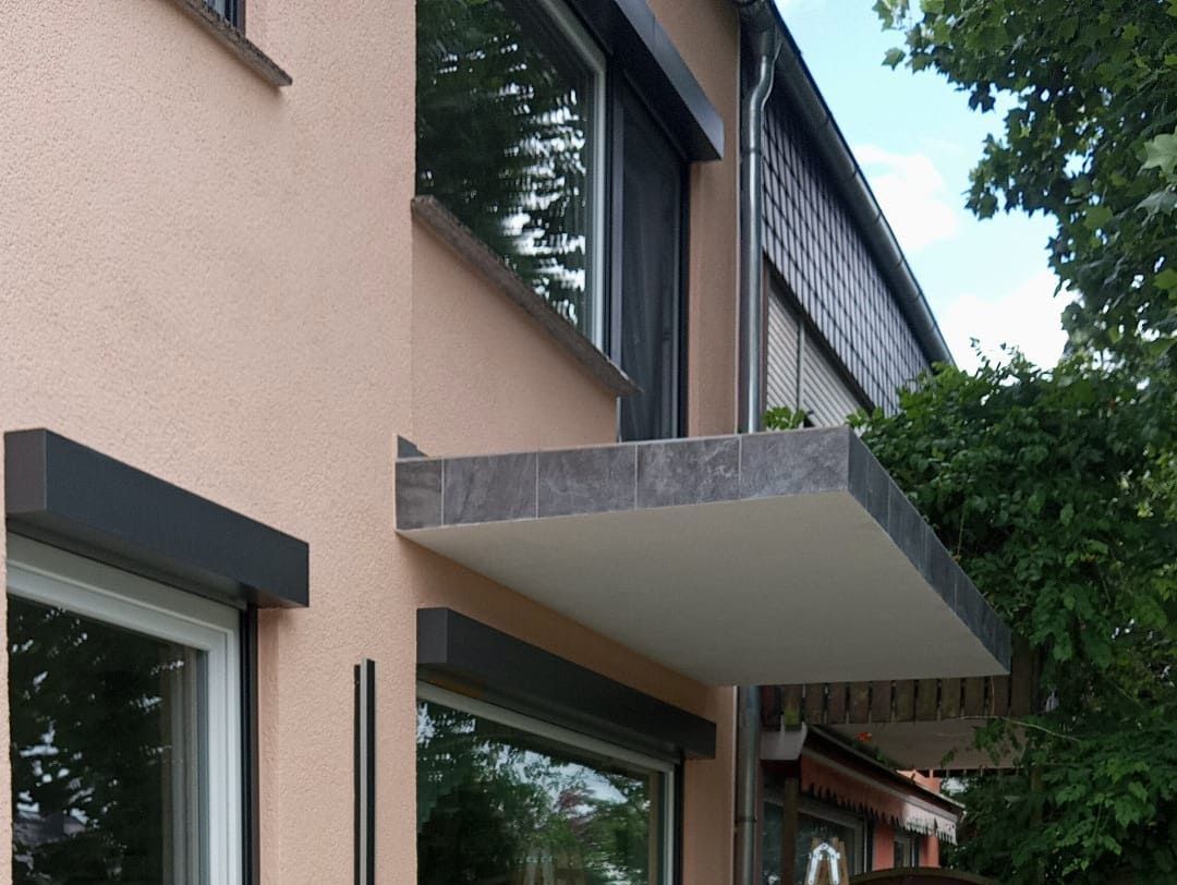 Außenansicht eines Gebäudes mit einer flachen, steingedeckten Markise. Dunkle Fensterrollos und rosafarbene Stuckwand.