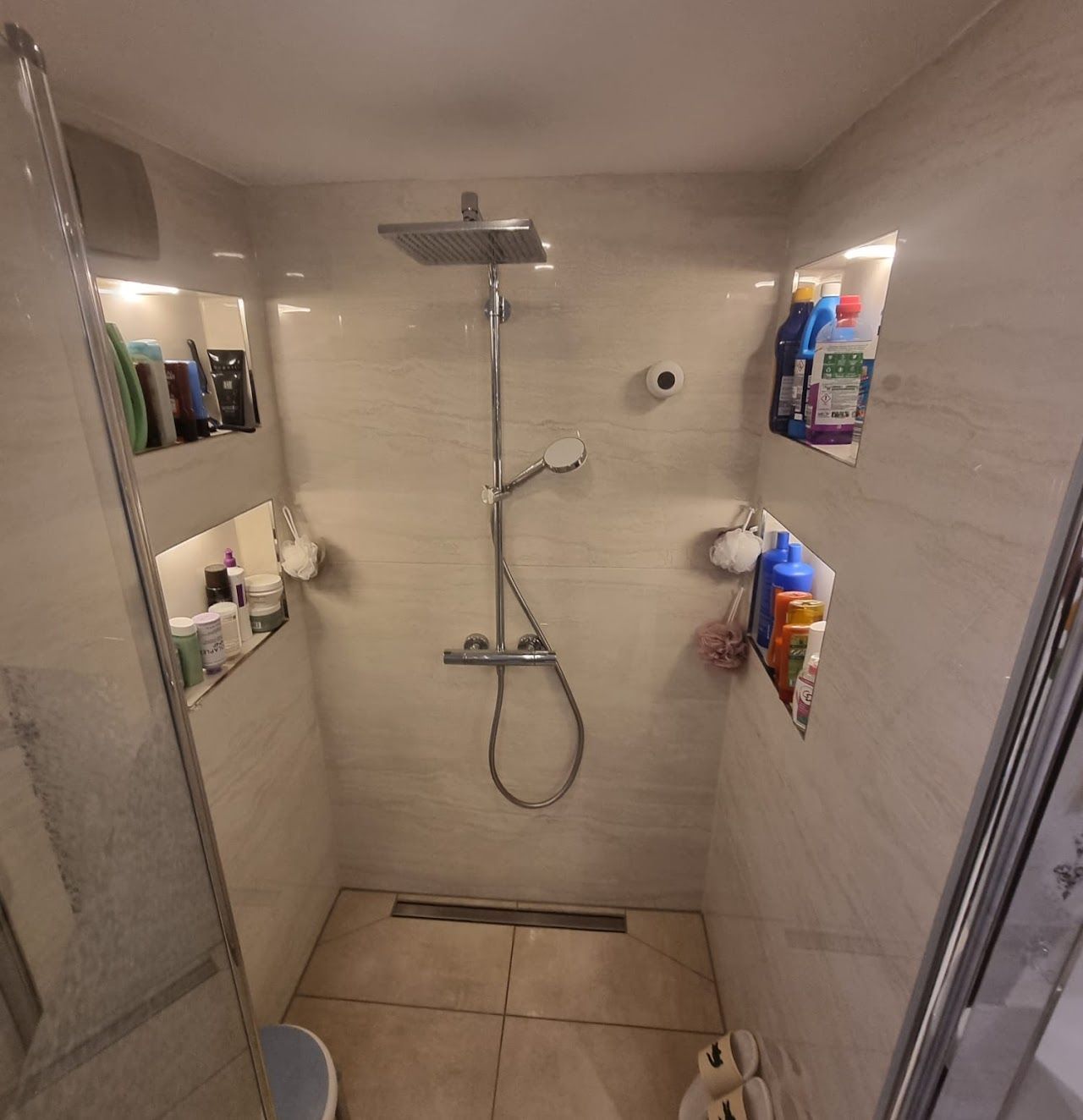 Duschkabine mit integrierten Ablagen für Toilettenartikel. Quadratischer Duschkopf, Handbrause und Glastür.