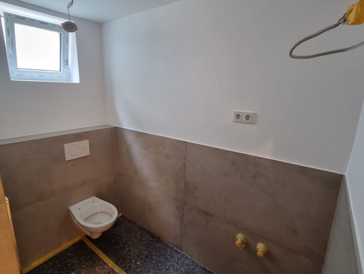 Badezimmer-Umbau: Ausgestattet mit Toilette, teilweise gefliesten Wänden, einem Fenster und freiliegenden Rohrleitungen.