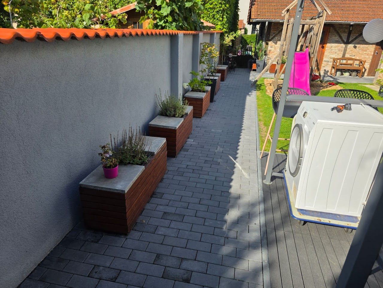 Pflanzkästen entlang eines grauen Backsteinwegs, neben einer grauen Mauer und einem Gebäude.