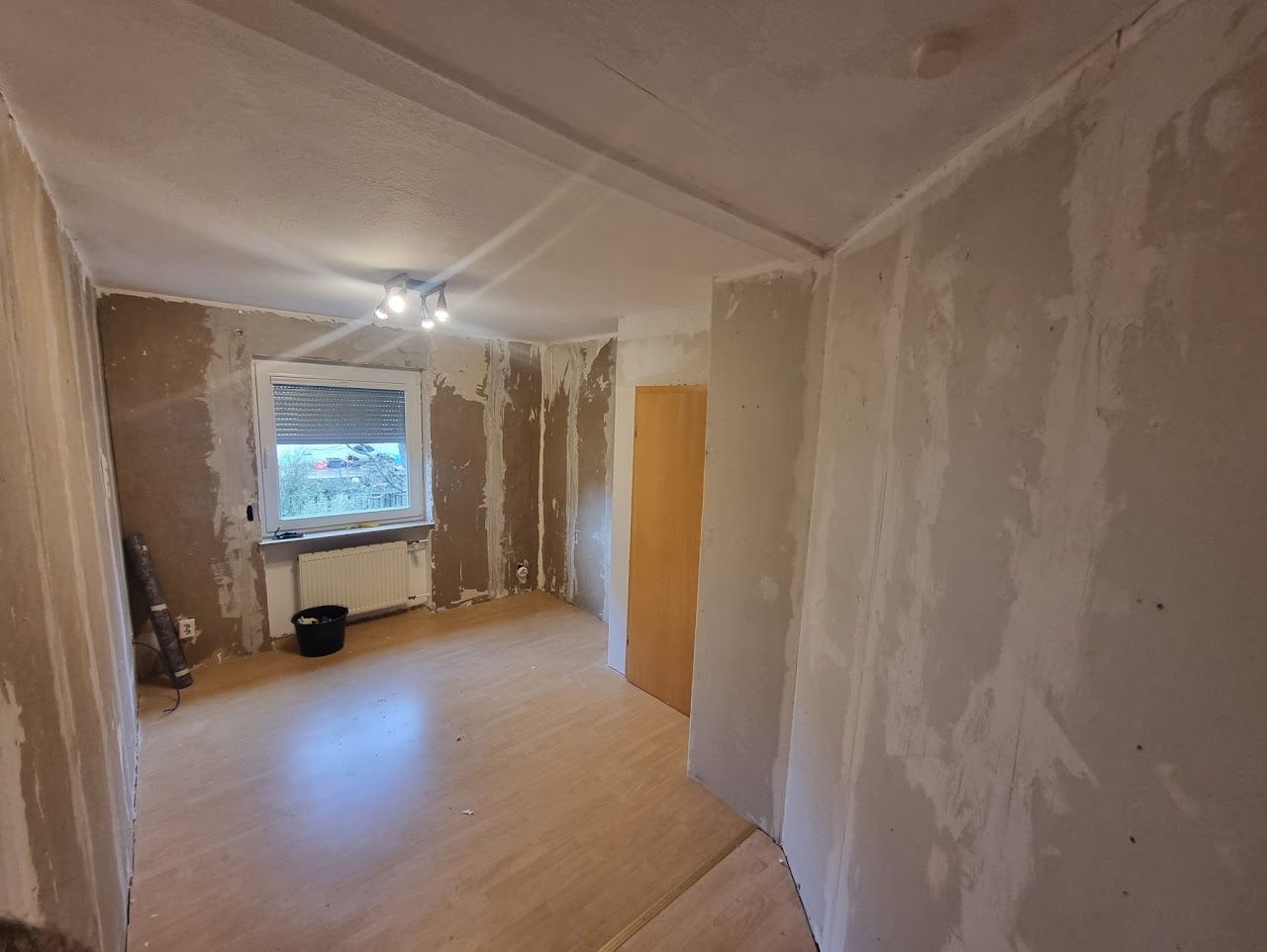 Zimmer im Umbau mit kahlen Wänden, Fenster, Holzboden und geschlossener Tür.