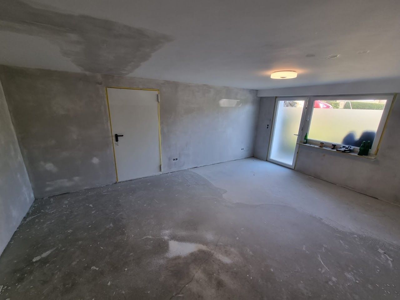 Zimmer im Umbau mit nackten Betonwänden, Betonboden sowie geschlossener Tür und Fenster.