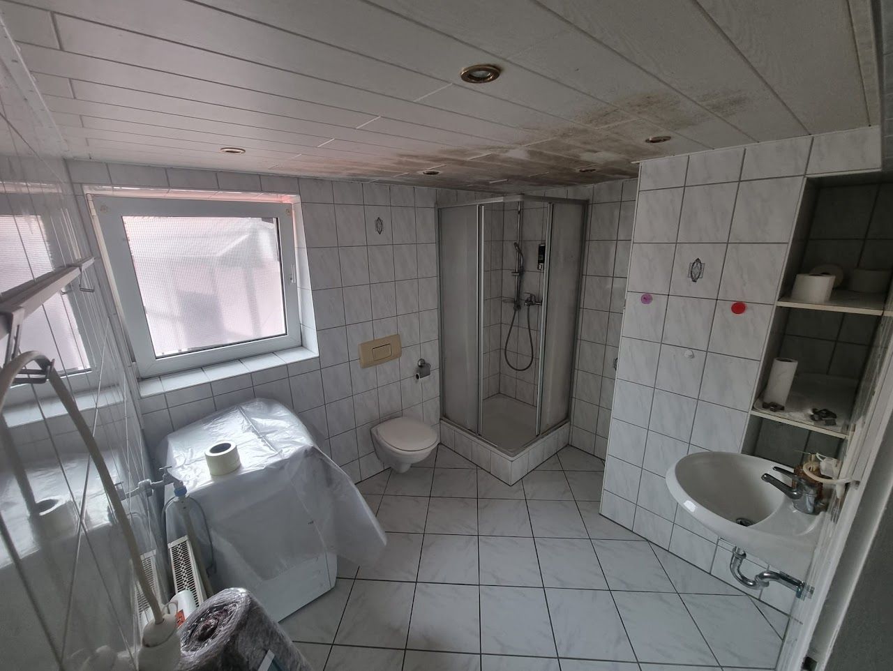 Badezimmer mit weißen Fliesen, Duschkabine, Toilette und Waschbecken. Die Decke weist Wasserschäden auf.