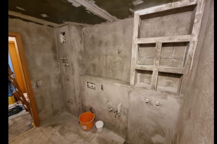 Badezimmer im Bau: Betonwände, unfertige Regale, freiliegende Rohre und Steckdosen, Eimer auf dem Boden.