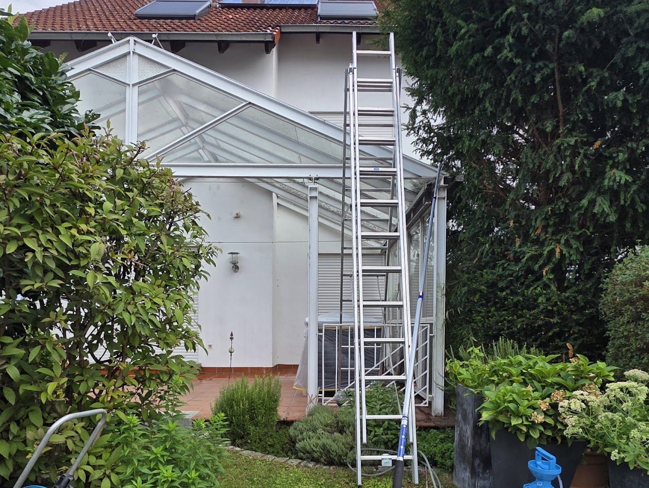 Eine Leiter lehnt an einer gläsernen Dachkonstruktion, die an ein weißes Gebäude angebaut ist; grünes Laubwerk umgibt sie.