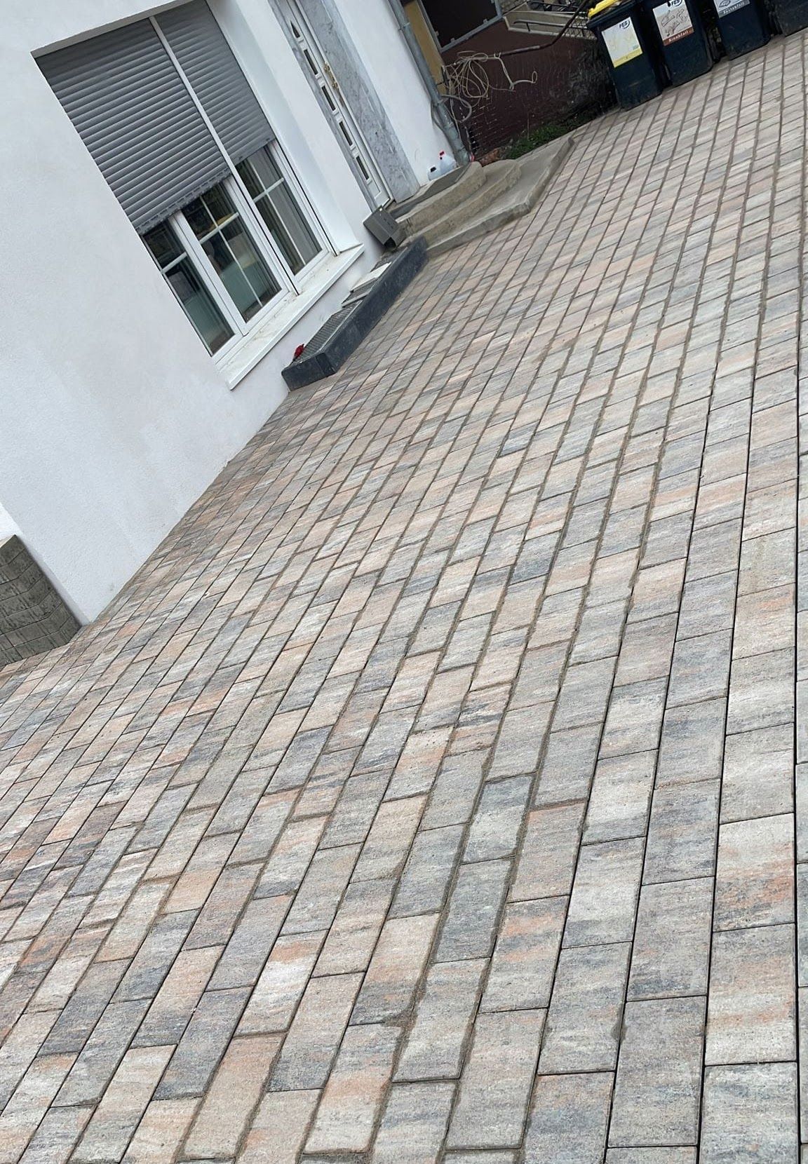 Eine mit Ziegeln gepflasterte Auffahrt führt an einem weißen Gebäude mit Fenster und Jalousien vorbei.