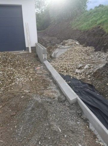 Baustelle: Betonentwässerungsrinne neben einem weißen Gebäude mit Garagentor und Kiesboden.