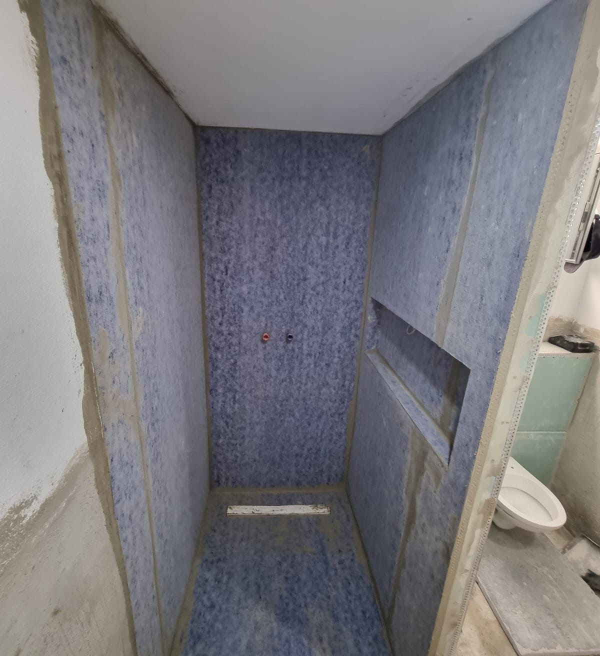 Duschkabine im Bau, blaue wasserdichte Paneele, rechteckige Nische, weiße Toilette sichtbar.