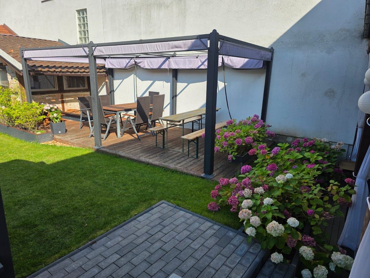 Hinterhofterrasse mit Pergola, Sitzgelegenheiten, umgeben von Rasen und blühenden Sträuchern.