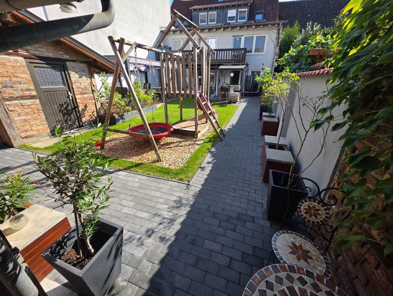 Hinterhof mit Spielplatz, Ziegelweg und Sitzgelegenheiten. Üppiger grüner Rasen und alte Bäume.