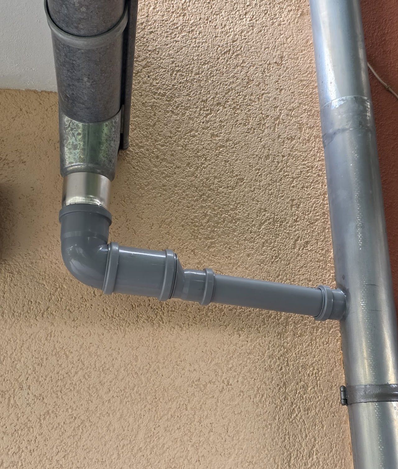 Graue und silberne Rohre sind an einer strukturierten Wand miteinander verbunden und bilden ein Entwässerungssystem.