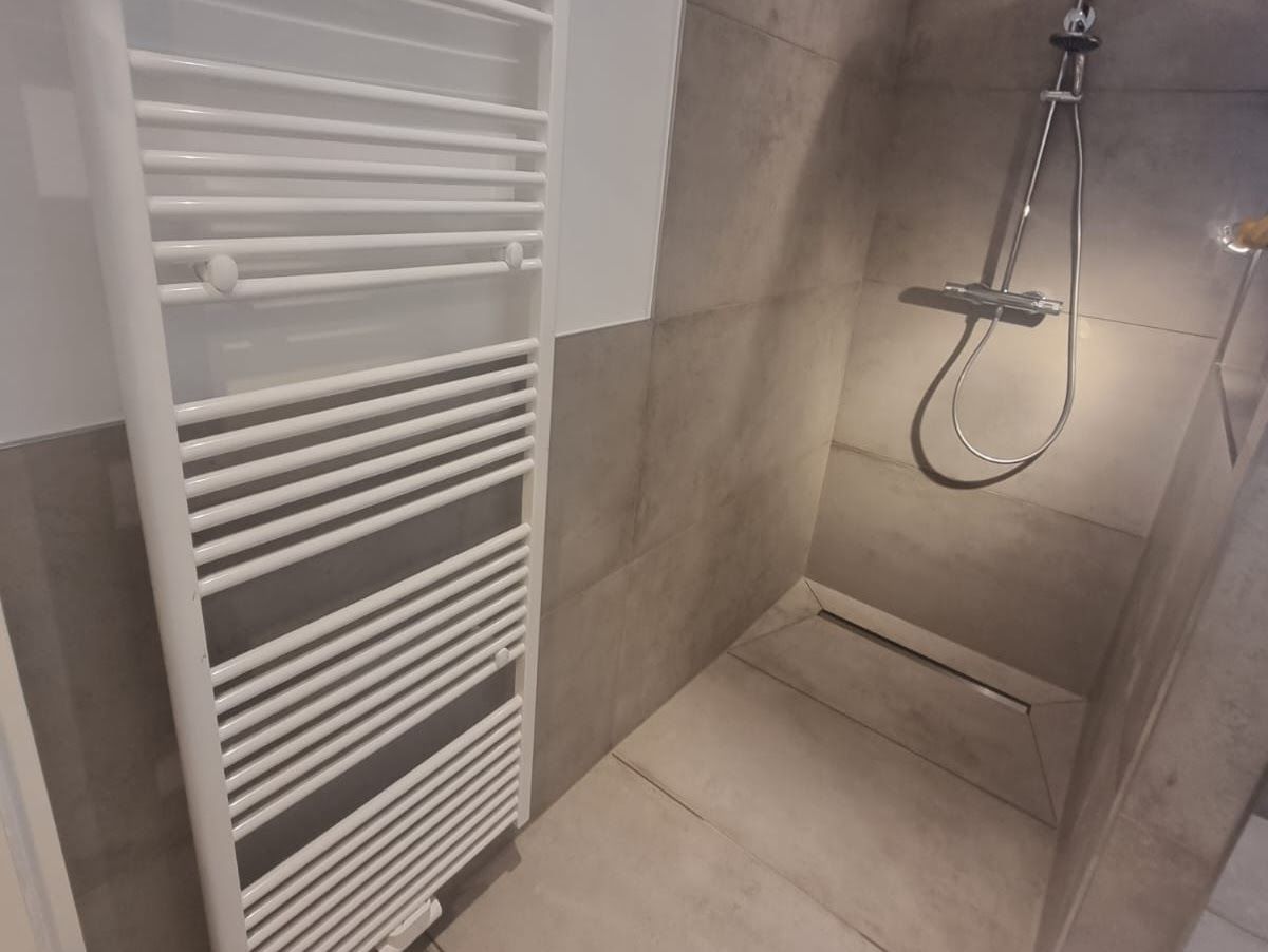 Ein weißer Heizkörper steht neben einer begehbaren Dusche mit grauen Fliesen und einem Duschkopf aus Chrom.