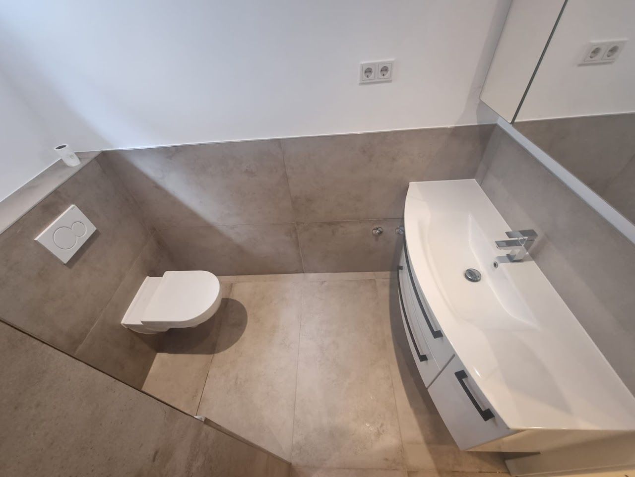 Modernes Badezimmer mit weißer Toilette, Waschbecken und grau gefliesten Wänden.