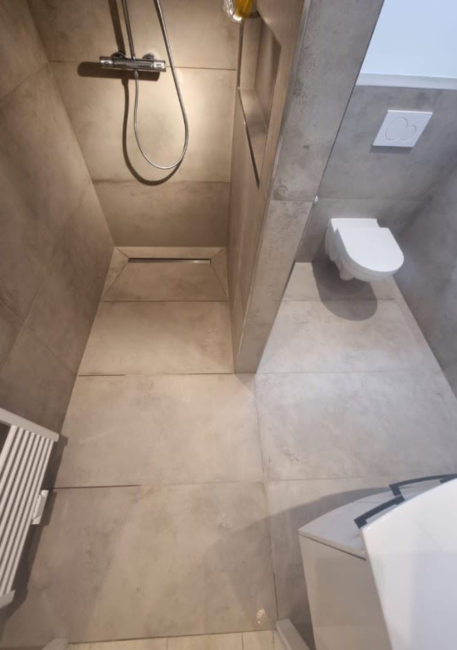 Ein modernes Badezimmer mit grauen Fliesen, Dusche, Toilette und einem kleinen weißen Heizkörper.