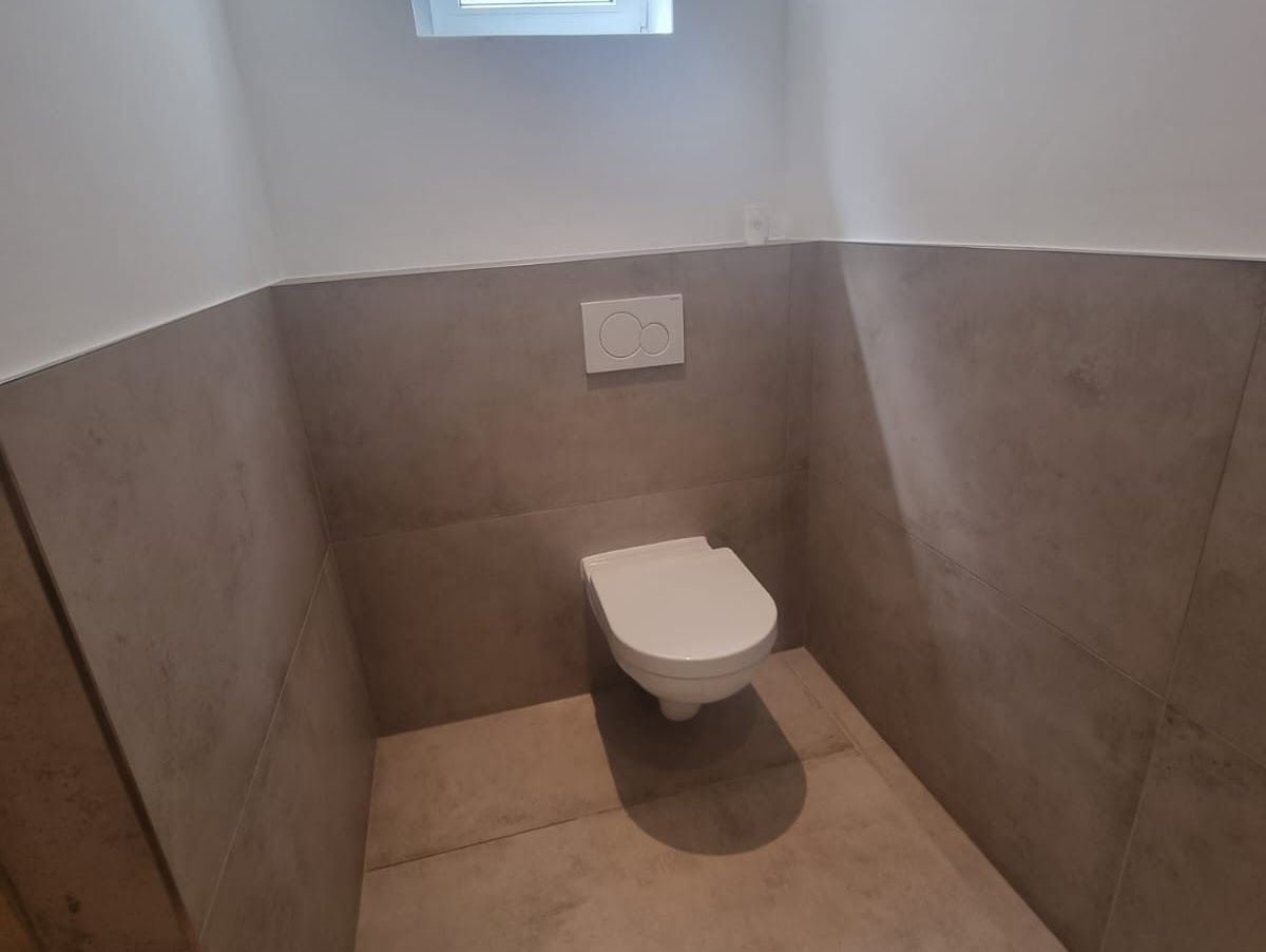 Toilette in einem kleinen Badezimmer. Beige geflieste Wände und Boden, weiße Toilette, Spülknopf, kleines Fenster darüber.