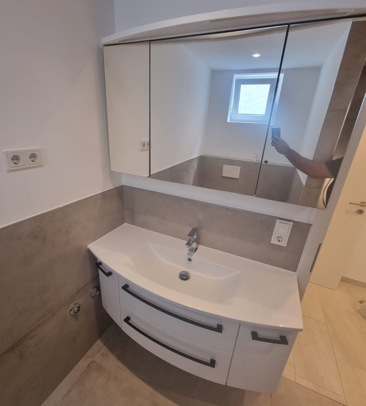 Badezimmer mit weißem Waschtisch, großem Spiegelschrank und einem kleinen Fenster.