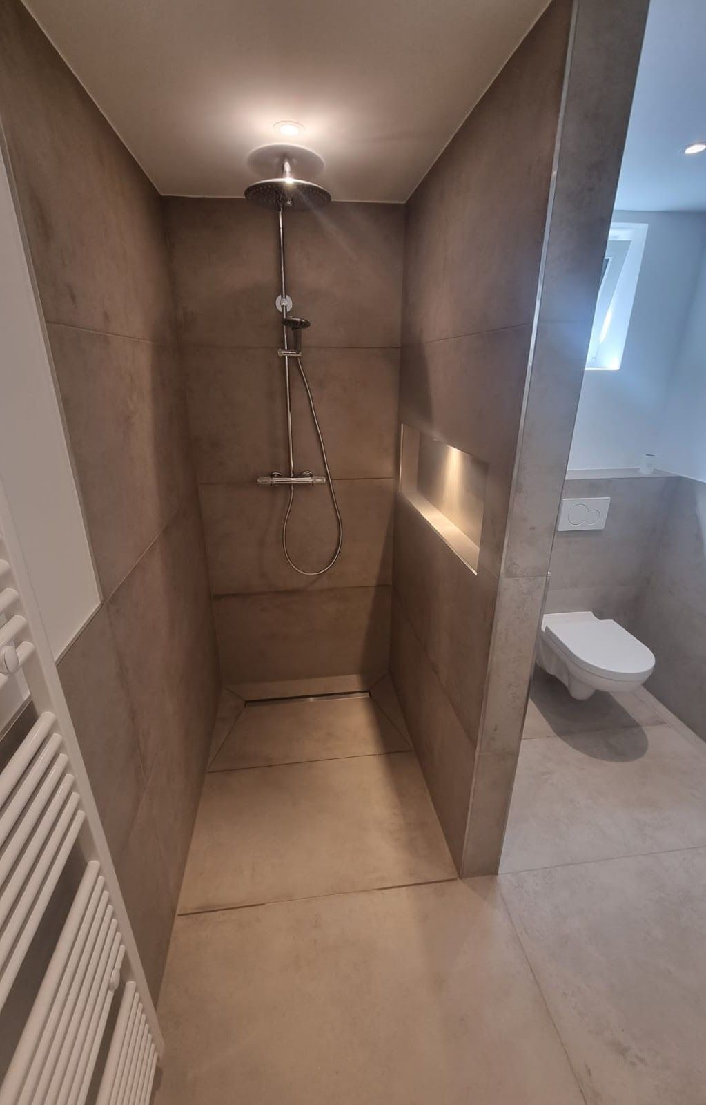 Eine moderne Duschkabine mit Regendusche und integriertem Regal. Beige Fliesenwände, Toilette sichtbar.