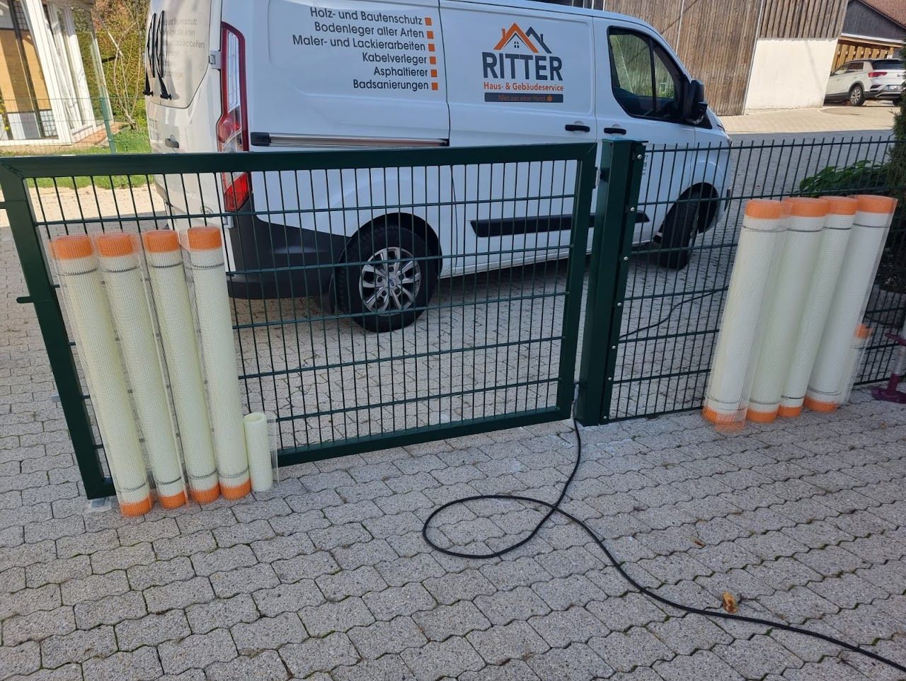 Grüner Metallzaun mit orangefarben ummantelter, gerollter Isolierung, die daran lehnt, vor einem weißen Lieferwagen.