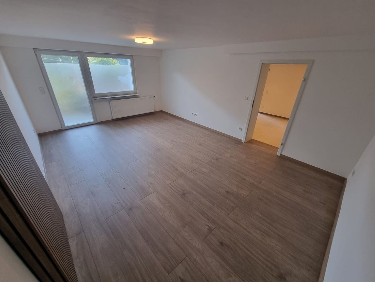 Leeres Zimmer mit Holzbodenoptik, weißen Wänden und einer Glasschiebetür.