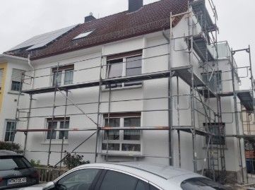 Hausfassade mit Renovierungsgerüst. Weiße Wände, rotes Dach, Solaranlage. Autos parken davor.