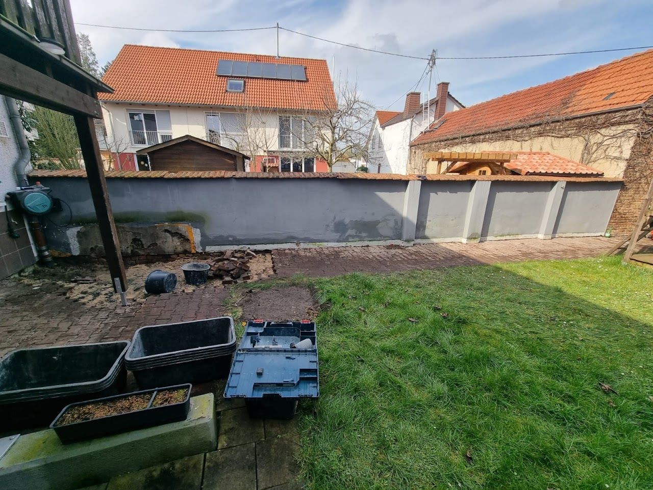 Hinterhof mit grauer Mauer, Gebäuden mit roten Dächern und Rasen. Mehrere Pflanzkübel stehen auf dem Boden.