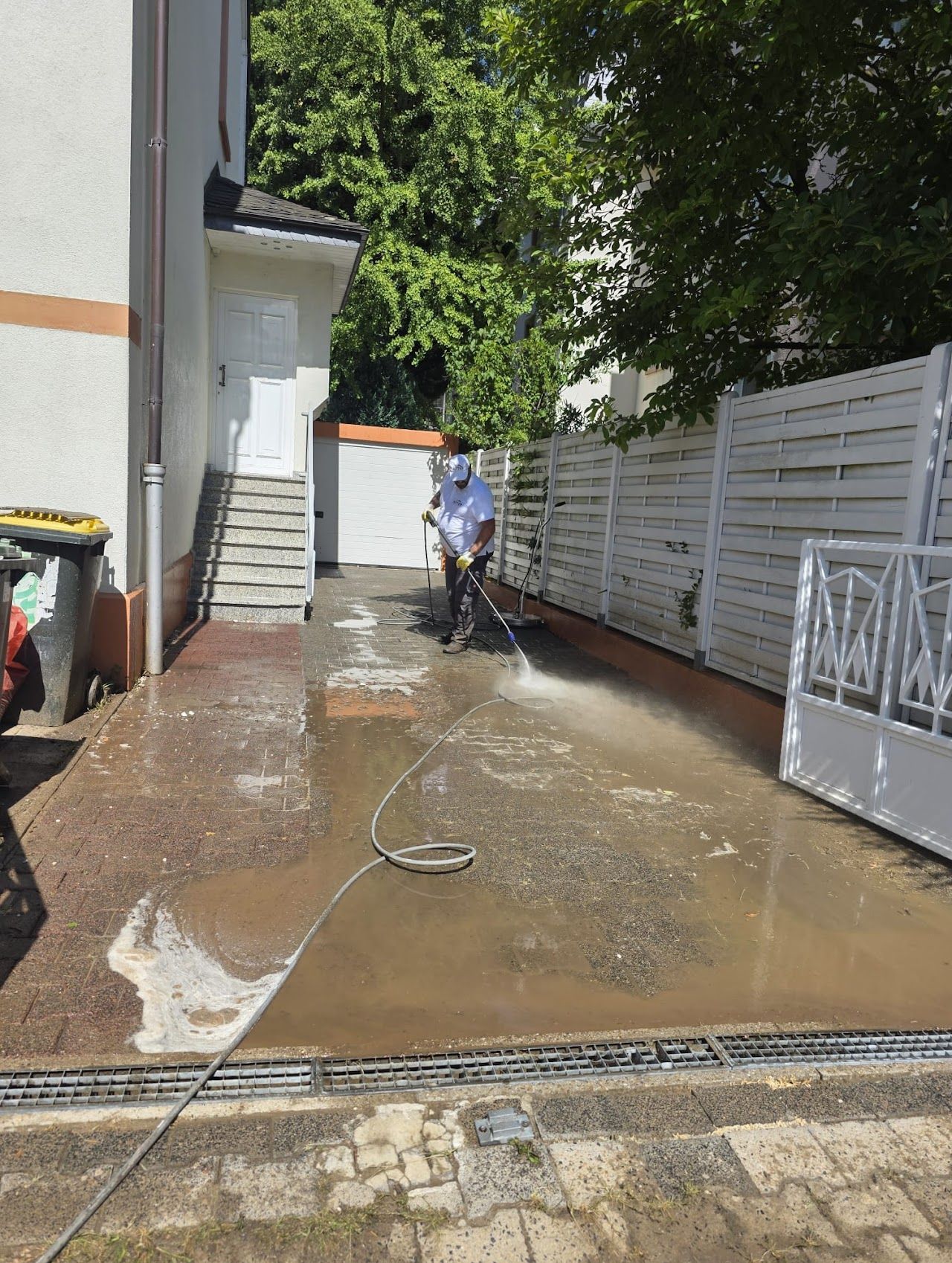 Eine Person reinigt mit einem Hochdruckreiniger eine gepflasterte Fläche neben einem weißen Zaun und einem Gebäude.