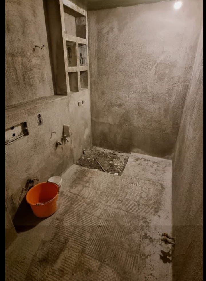 Badezimmer im Bau mit rauen Betonwänden und -boden. Ein orangefarbener Eimer steht auf dem Boden.