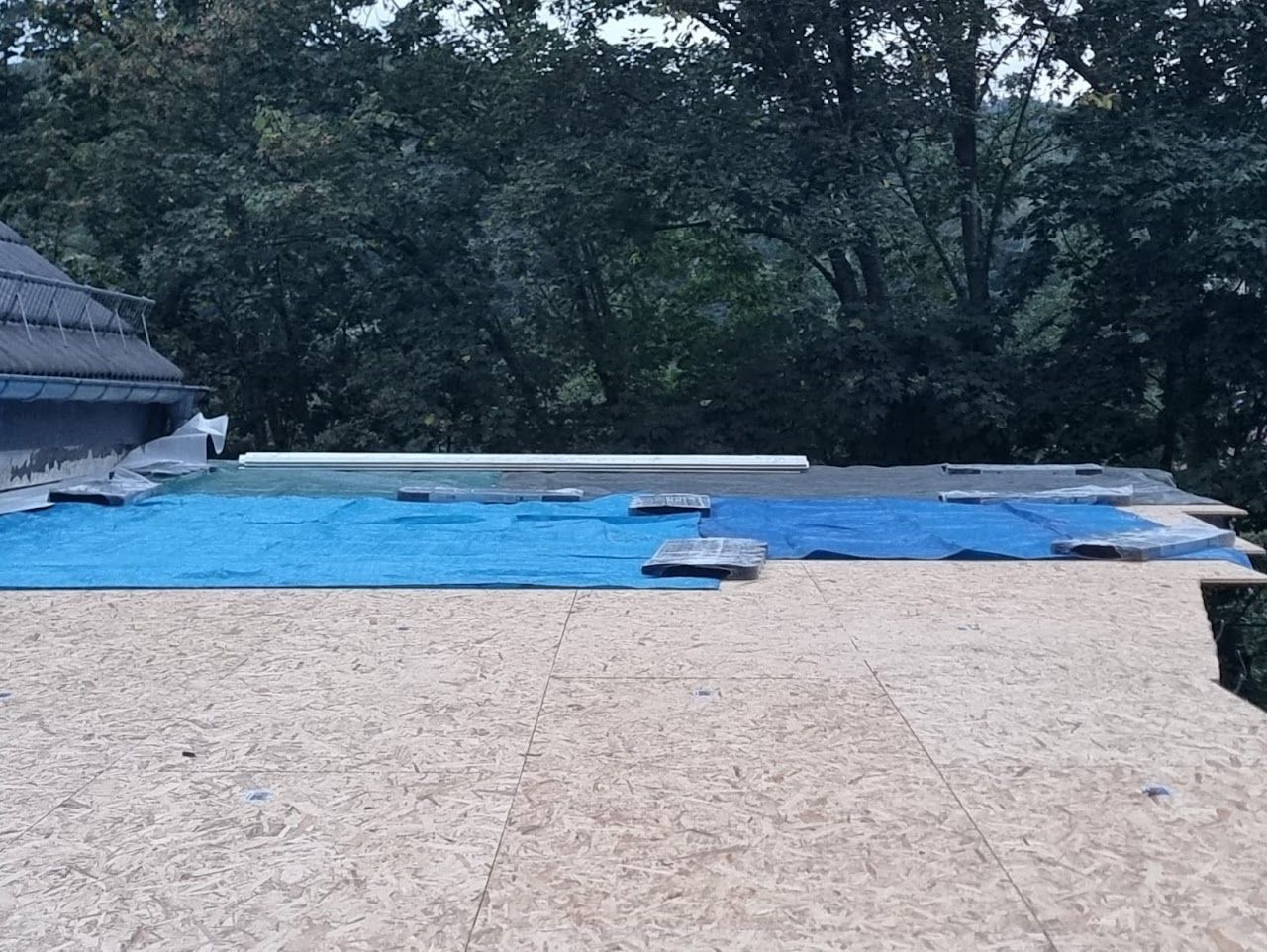 Dachterrasse mit blauer Plane und Holzbrettern, Bäume im Hintergrund.
