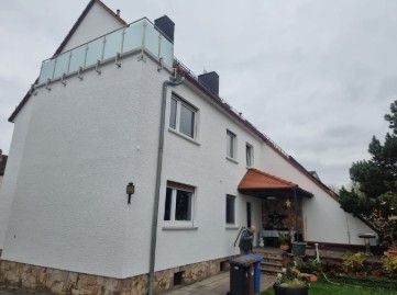 Weißes zweistöckiges Haus mit Glasbalkon, rotem Dach und Steinsockel an einem bewölkten Tag.