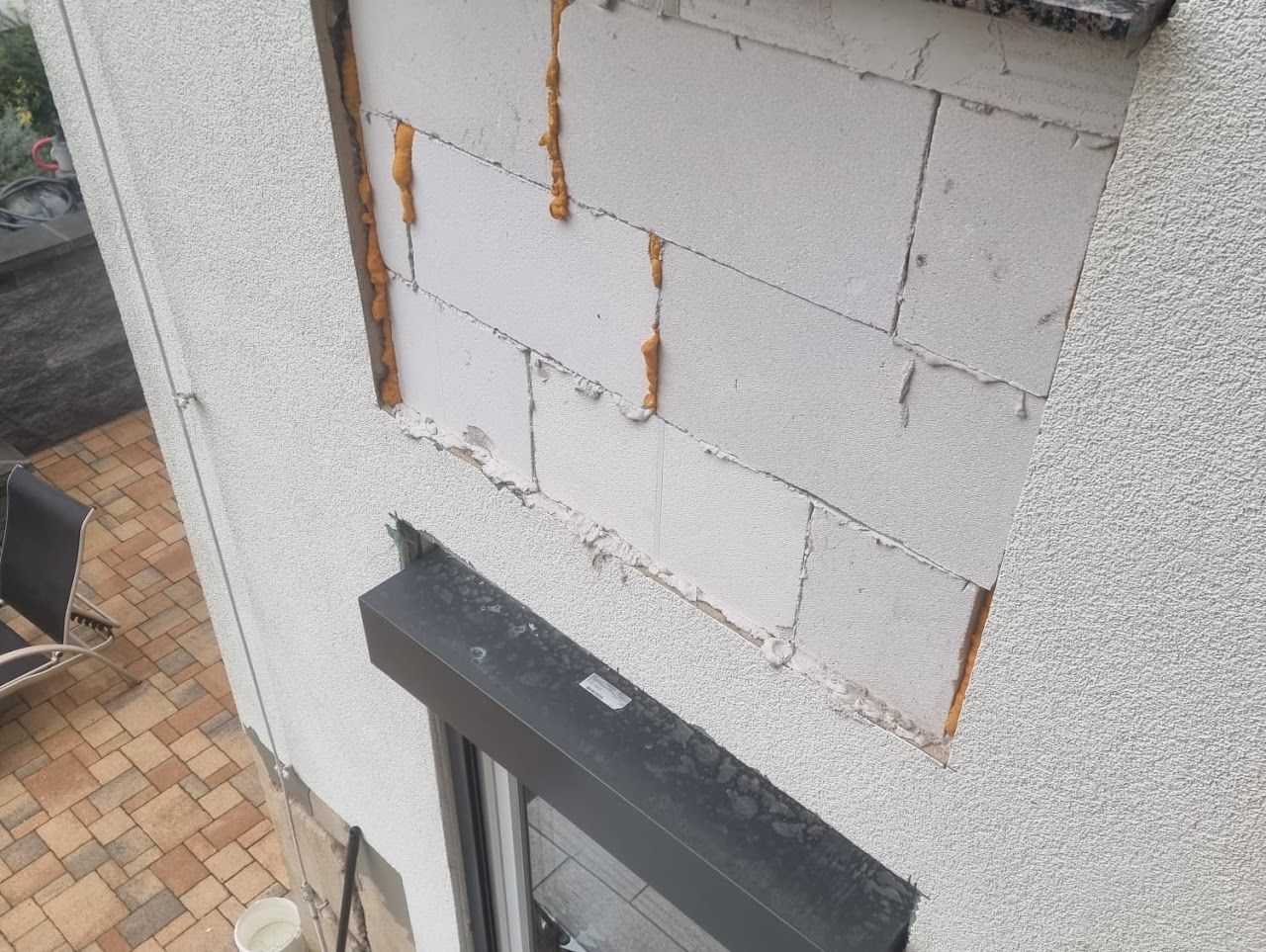 Außenwand mit freiliegendem weißen Ziegelstein, reparaturbedürftig. Schwarzer Fensterladenrahmen unten.