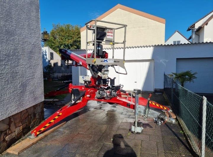 Rote Gelenkhebebühne auf einer gepflasterten Fläche, Arme ausgefahren, in der Nähe eines Gebäudes und einer Garage.