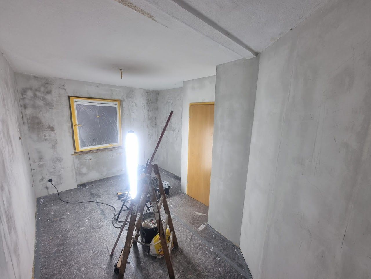 Ein Raum im Umbau mit hellem Arbeitslicht. Die Wände sind grau verputzt, das Fenster ist gelb umrandet.