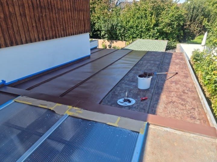 Flachdach, auf dem braunes Dachmaterial verlegt wird; Eimer und Werkzeug sind vorhanden.