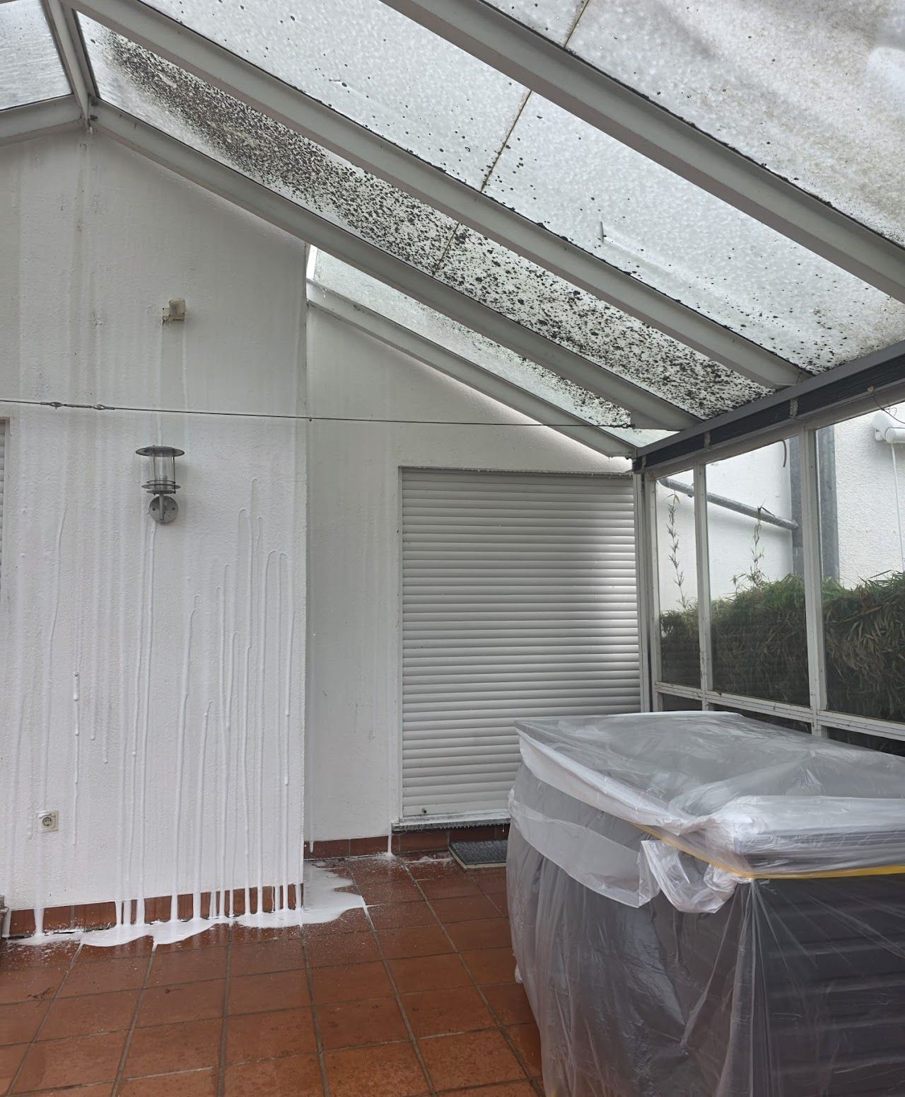 Eine überdachte Terrasse mit weißer Wand, transparentem Dach und einem mit Plastikfolie abgedeckten Tisch.