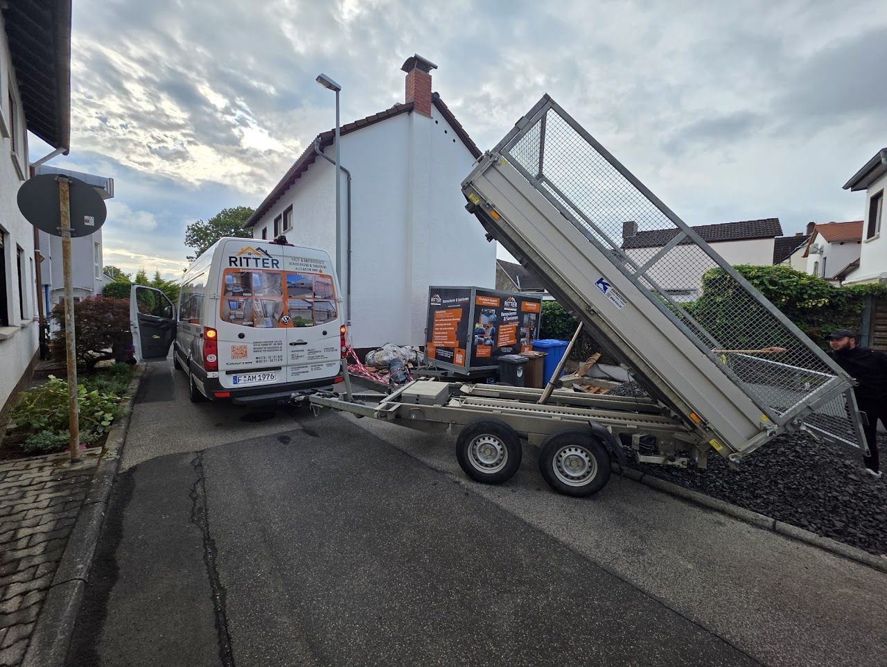 Ein Lieferwagen mit Anhänger kippt Kies in einer schmalen Wohnstraße ab. Ein weißes Gebäude ist zu sehen.