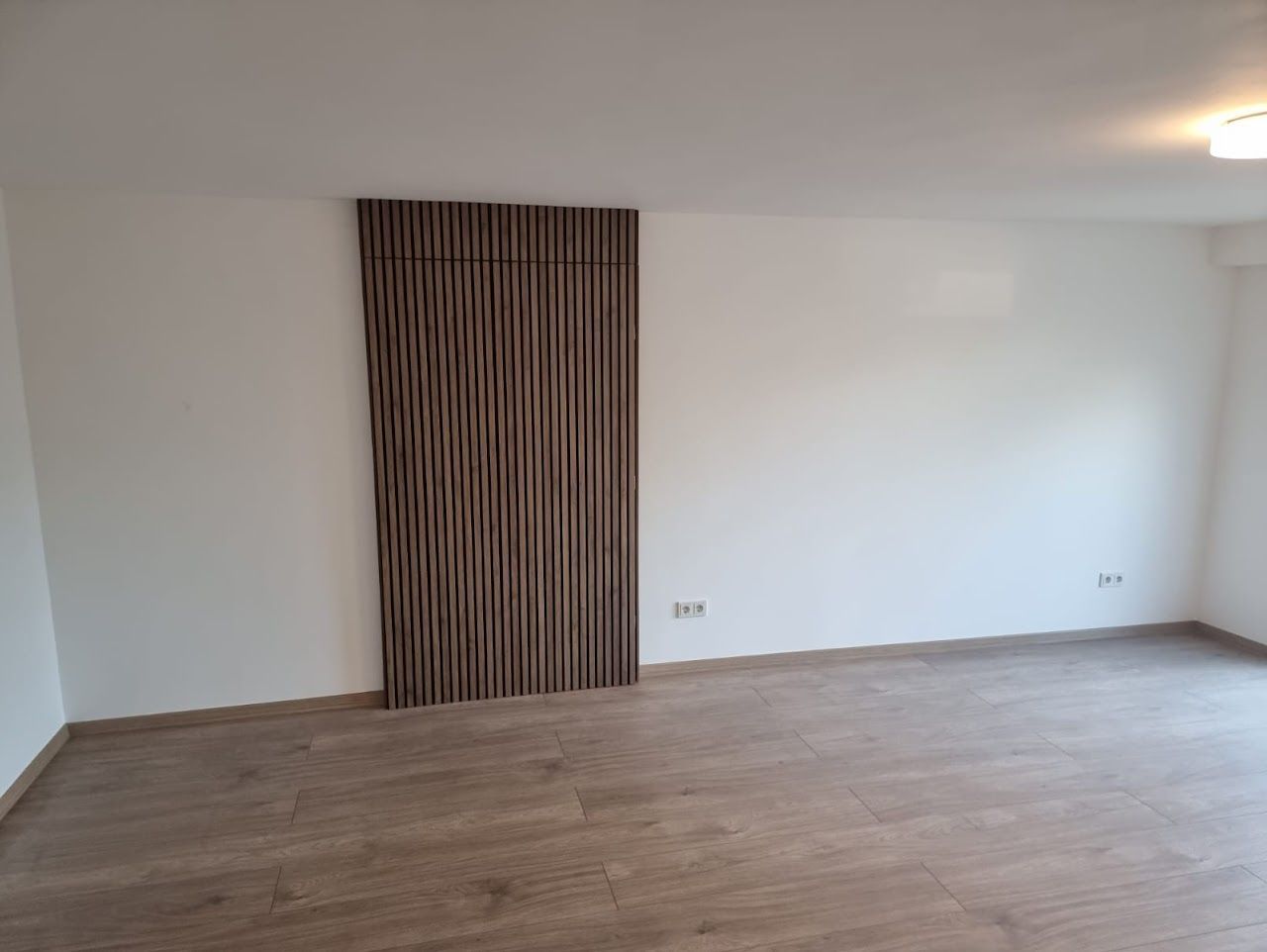 Leeres Zimmer mit holzstrukturierter Wandverkleidung, hellbraunem Boden und weißen Wänden.