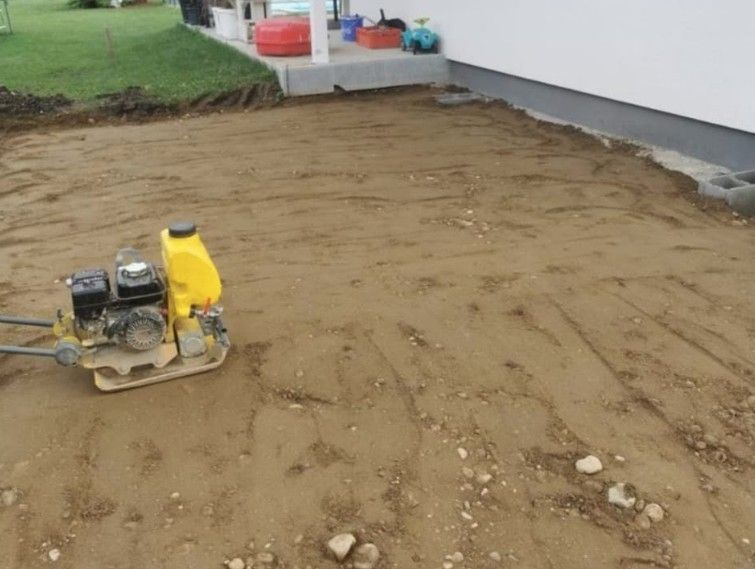 Eine gelbe Rüttelplatte verdichtet den Boden in einem Wohngarten in der Nähe eines Hausfundaments.