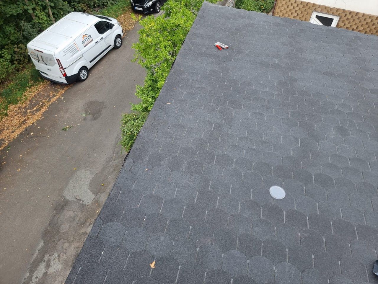 Blick vom Dach auf eine Straße mit Asphaltziegeln, einem weißen Lieferwagen, einem schwarzen Auto und Bäumen.