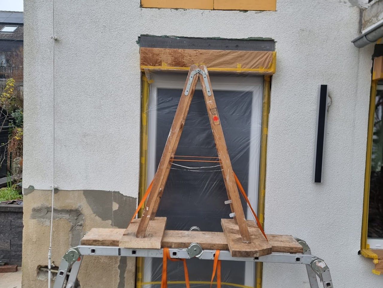 Hölzerne Stützkonstruktion auf einer Leiter vor einem Fenster während Bauarbeiten an der Gebäudeaußenfassade.