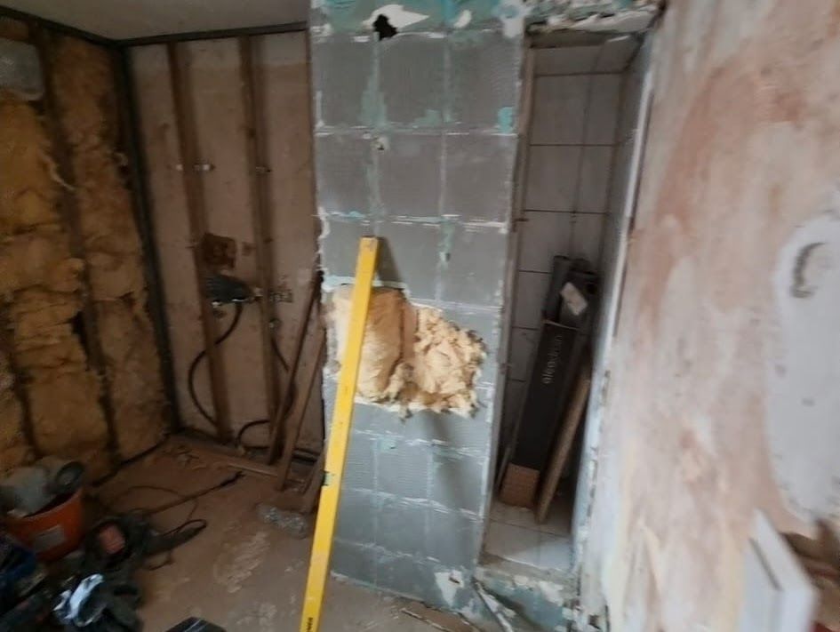 Die Innenrenovierung ist im Gange, beschädigte Wand mit freiliegender Dämmung und Fliesen; gelbe Wasserwaage dagegen gelehnt.