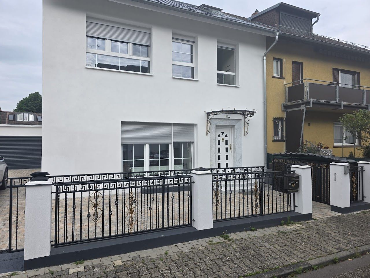 Weißes Haus mit schwarzem Zaun, sichtbaren Fenstern und einem gelben Haus nebenan.