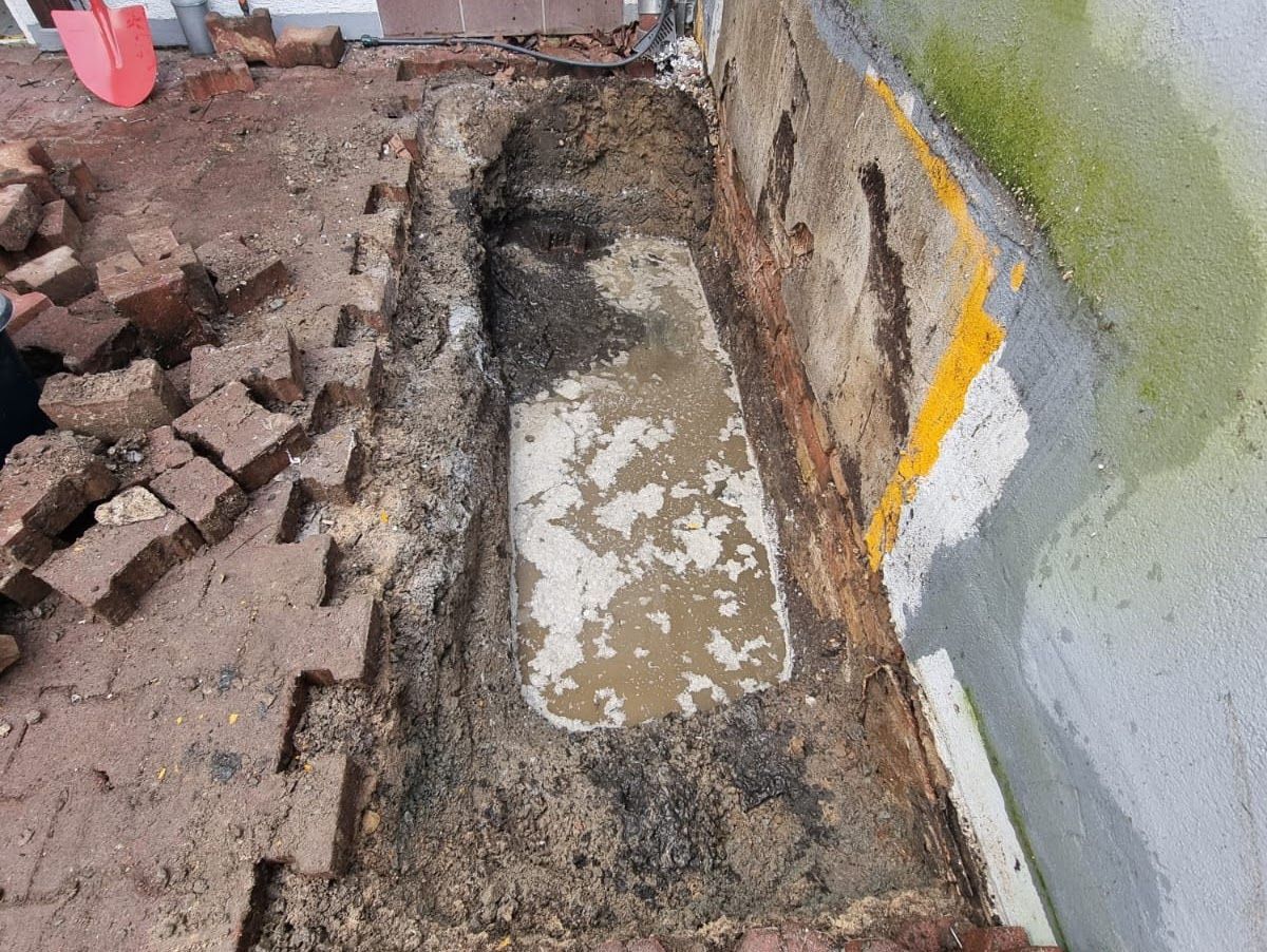 Ausgehobene rechteckige Grube mit Wasser in einer Mauer aus Ziegeln und Beton. Ziegelsteine ​​sind in der Nähe aufgestapelt.