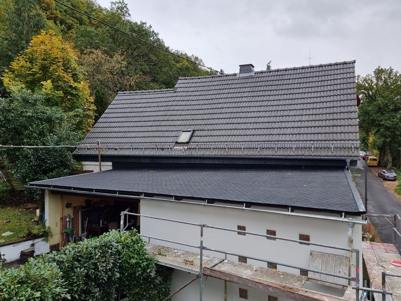 Ein Haus mit einem dunkelgrauen Ziegeldach und einem schwarzen Flachdach vor dem Hintergrund grüner Bäume.
