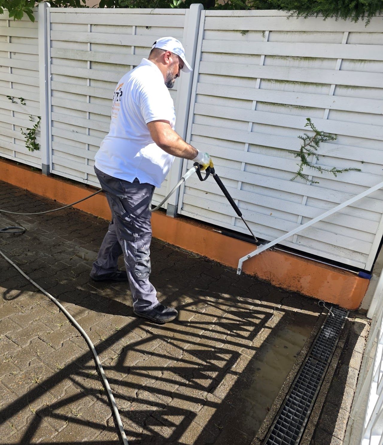 Ein Mann reinigt mit einem Hochdruckreiniger einen weißen Zaun und eine Ziegelterrasse.
