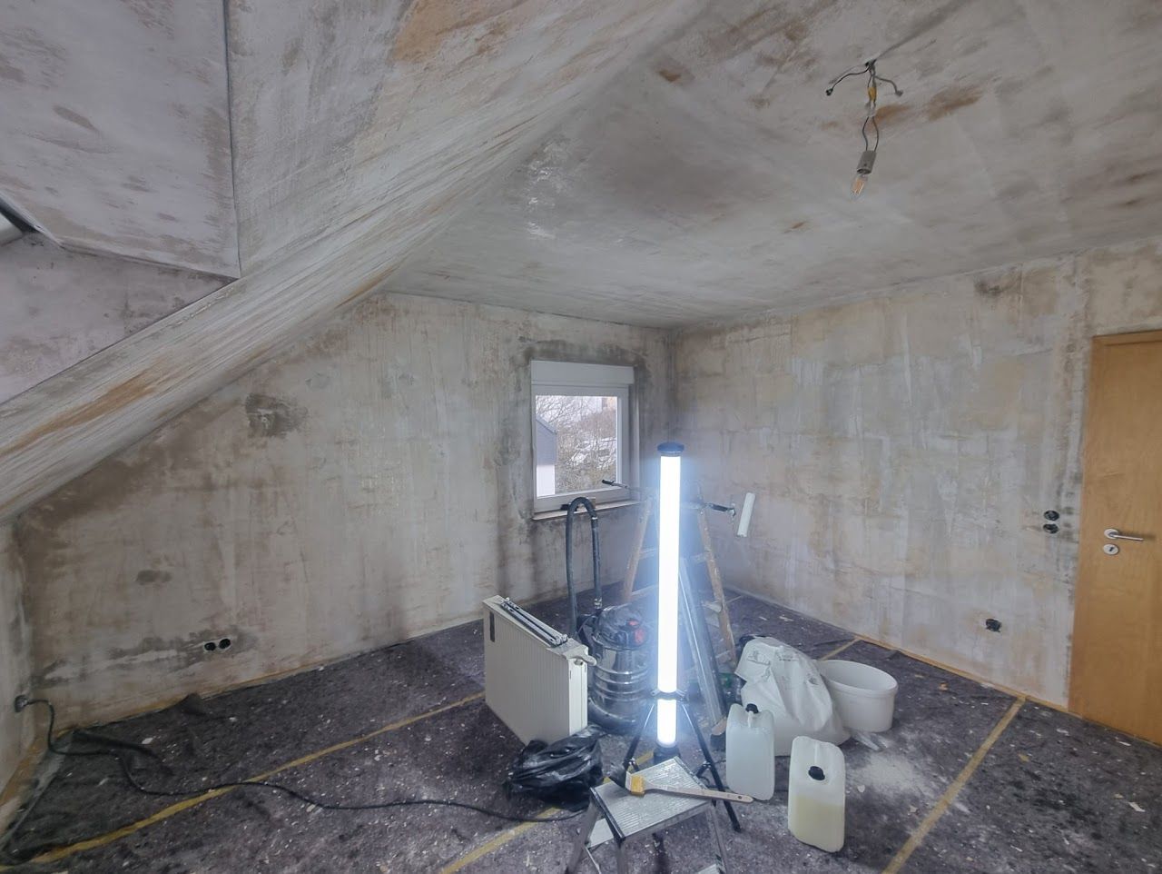 Ein Raum, der gerade renoviert wird, mit strukturierten Wänden und Boden, Baumaterialien und einer Arbeitsleuchte.