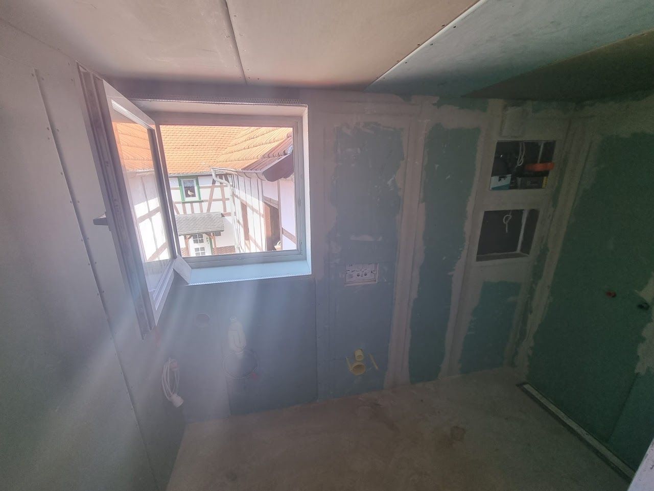 Innenraum im Bau, mit freiliegenden Gipskartonwänden, einem Fenster und Elektroverteilerkästen.
