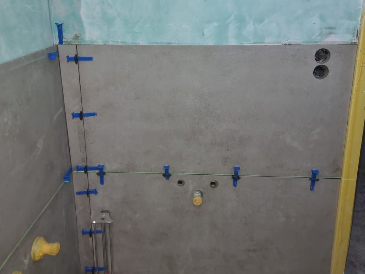 Badezimmerwand, graue Zementplatten mit blauen Fliesenverstellklammern.